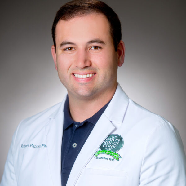 A man in a white lab coat with a badge stands against a plain background and smiles at the camera.