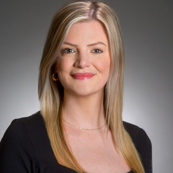 Smiling woman with straight blonde hair wearing a black top, gold necklace, and hoop earrings against a gray background.