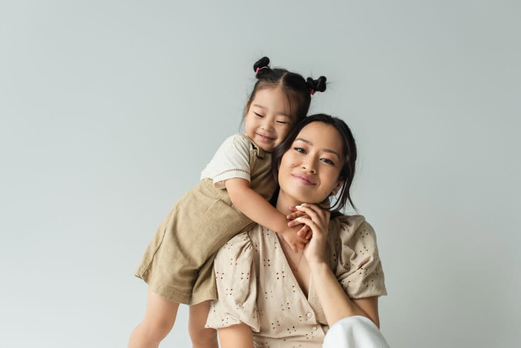A young girl with pigtails hugs a seated woman from behind. Both are smiling and wearing light-colored clothing against a plain background.