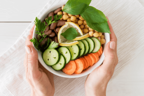 A person holds a bowl with sliced cucumber, carrot, chickpeas, kidney beans, herbs, and a lemon slice on a white cloth.