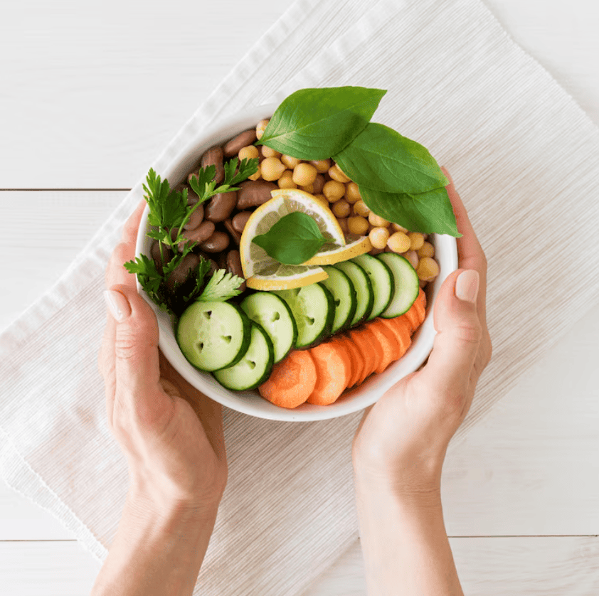 A person holds a bowl with sliced cucumber, carrot, chickpeas, kidney beans, herbs, and a lemon slice on a white cloth.