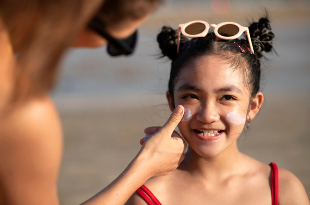 An adult applies sunscreen to a smiling girl’s face at the beach. The girl wears sunglasses on her head and a red swimsuit.
