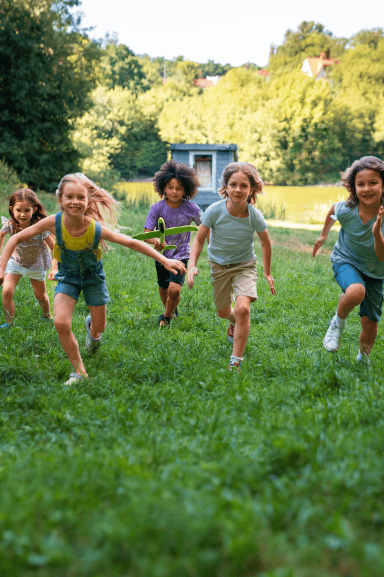 Five children run toward the camera across a grassy field in a park, with trees and a small building visible in the background.