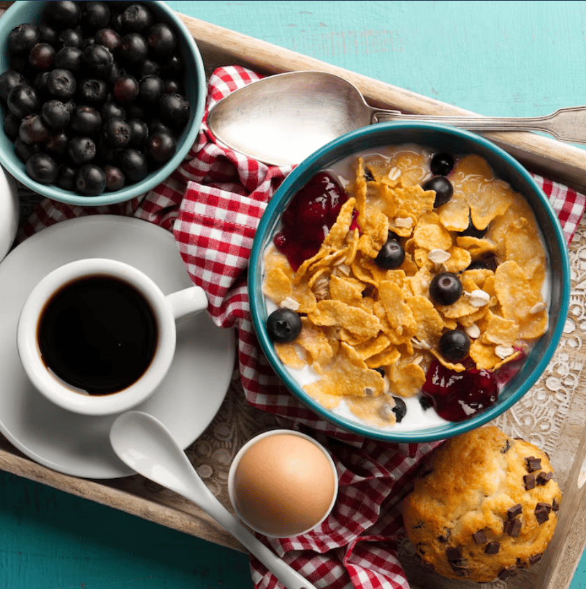 A tray with cereal and blueberries in milk, a boiled egg, a cup of coffee, a bowl of blueberries, and a chocolate chip muffin.