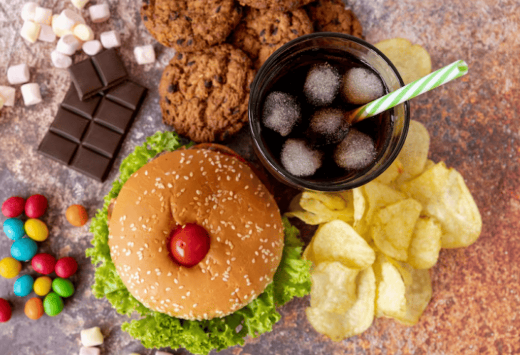 A hamburger, potato chips, chocolate, cookies, colorful candies, mini marshmallows, and a glass of soda with ice and a straw on a table.