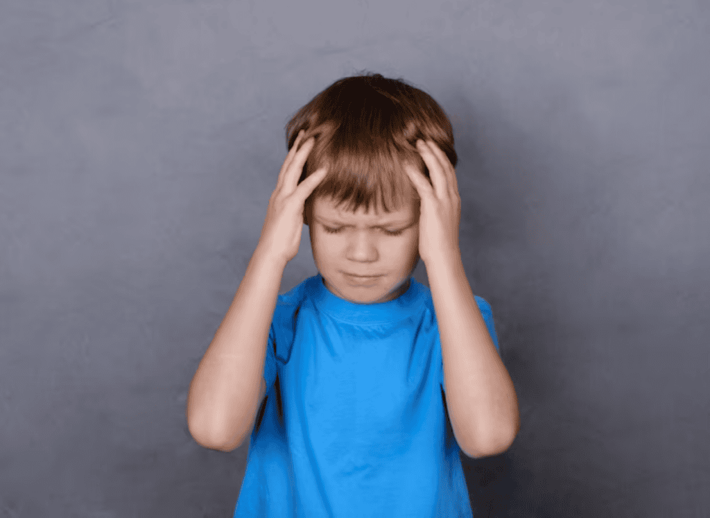 Young boy in a blue shirt holding his head with both hands, eyes closed, standing against a plain gray background.