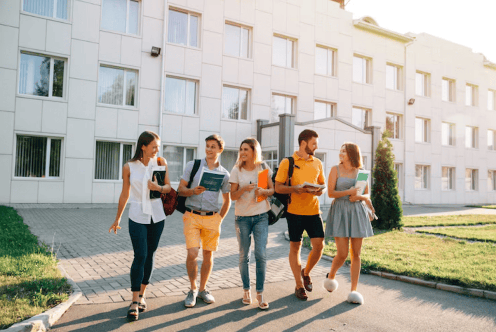 Five young adults walk together outside a modern building, carrying books and backpacks, appearing to be students.