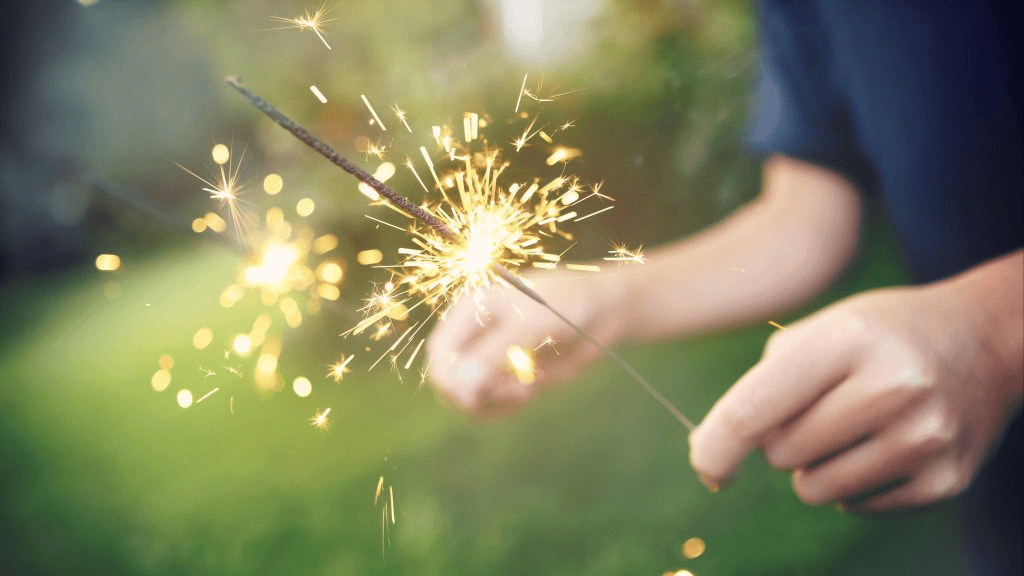 A person holds a lit sparkler outdoors, with sparks flying and a blurred green background visible.