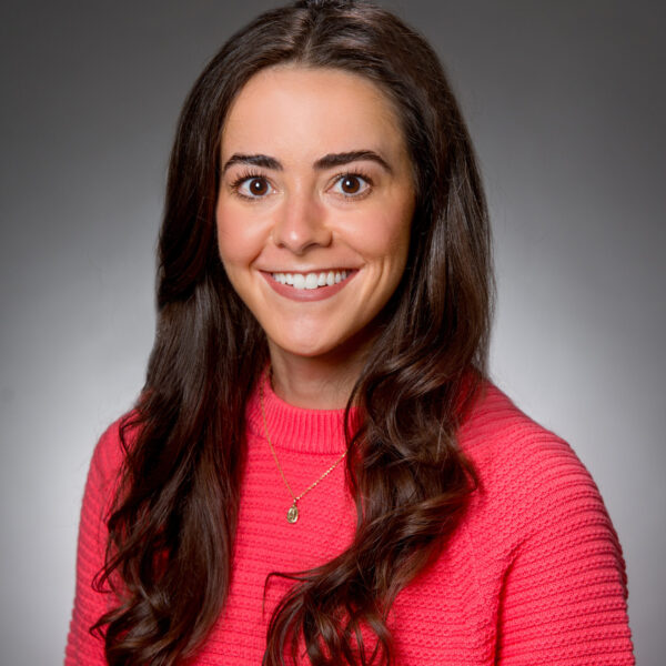 A woman with long brown hair wearing a bright pink textured sweater, smiling in front of a plain gray background.