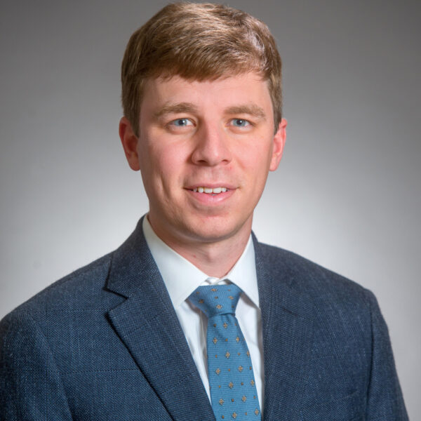 A man in a blue suit, white shirt, and blue patterned tie poses for a formal portrait against a plain gray background.