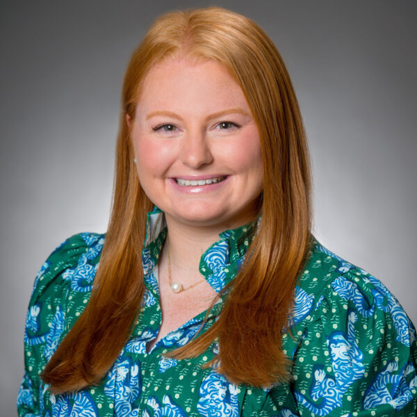 A woman with long red hair wearing a blue and green patterned blouse smiles at the camera against a gray background.
