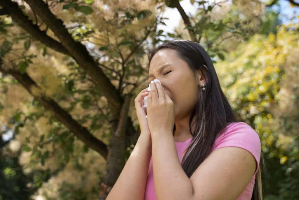 A person in a pink shirt sneezes into a tissue while standing outdoors near a tree on a sunny day.