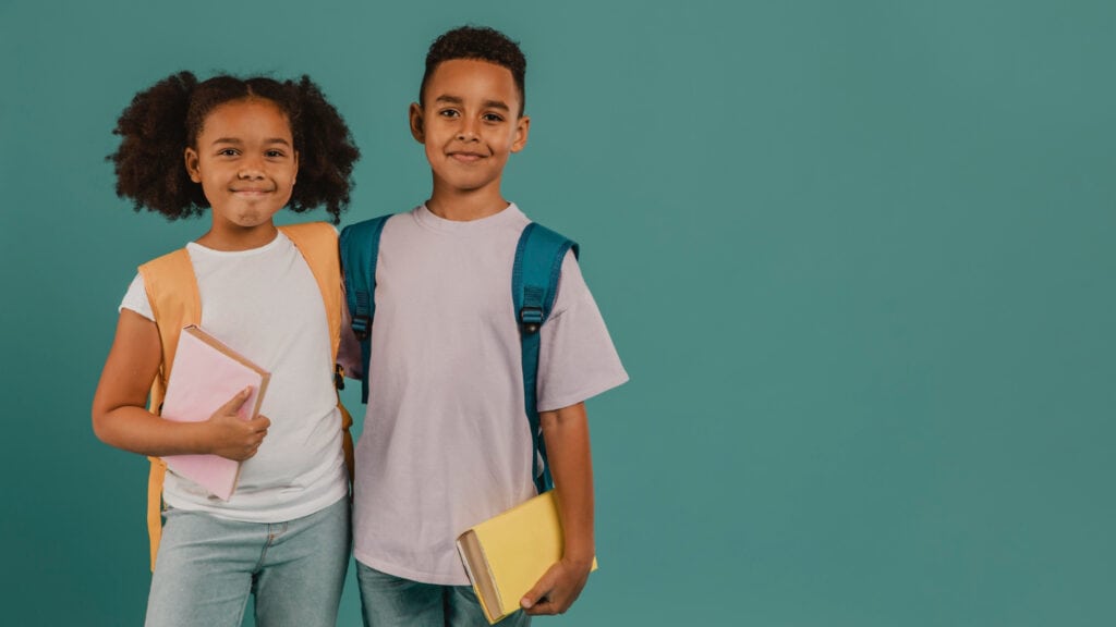 Two children with backpacks stand side by side against a teal background, each holding books and smiling at the camera.