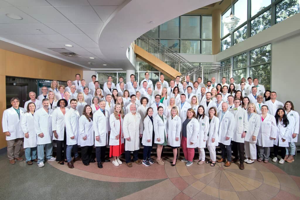 A large group in white lab coats gathers for a photo at Baton Rouge Clinic in a spacious, modern building with a staircase behind them.