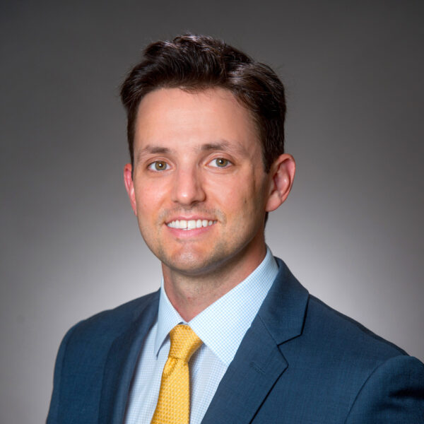 Man in a blue suit and yellow tie poses against a plain, gray background, facing the camera and smiling.