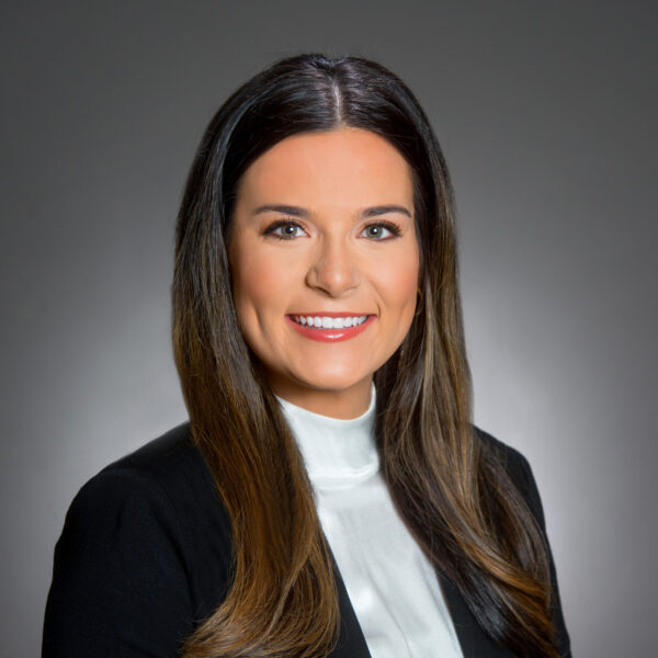 Woman with long brown hair wearing a black blazer and white blouse, smiling, posed in front of a plain gray background.