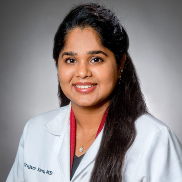 A woman with long dark hair wearing a white lab coat and a red top, smiling in a professional portrait against a gray background.
