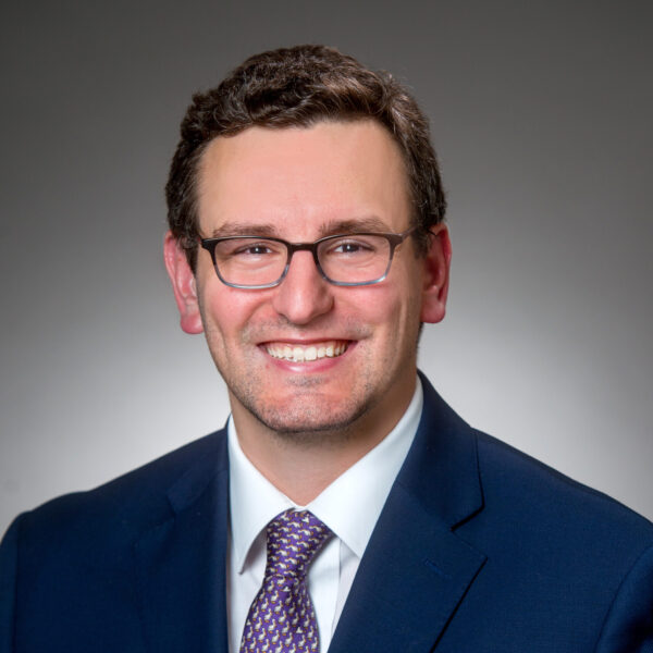 A man wearing glasses, a navy blue suit, a light shirt, and a patterned tie, smiling in front of a plain gray background.