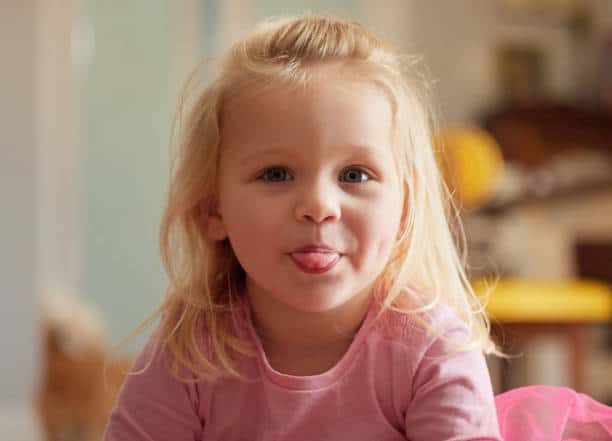 A young child with long blonde hair in a pink shirt sticks their tongue out, looking directly at the camera indoors.