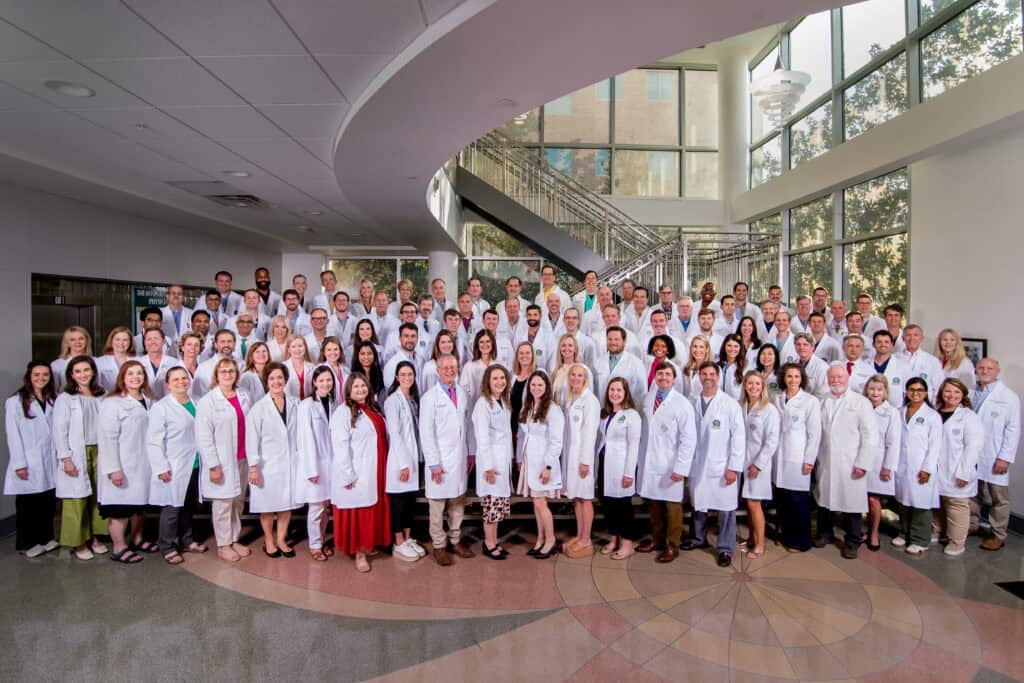 A large group in white lab coats pose for a group photo inside Baton Rouge Clinic, surrounded by large windows and a modern staircase.
