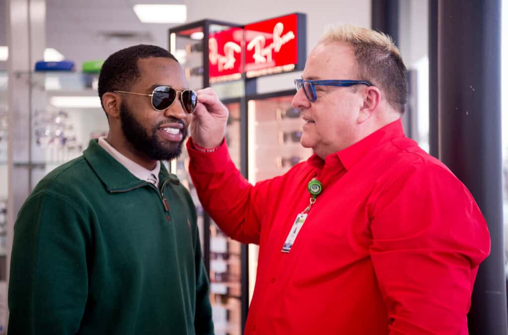 A man in a red shirt helps another man try on sunglasses in an eyewear store. Glasses displays are visible in the background.