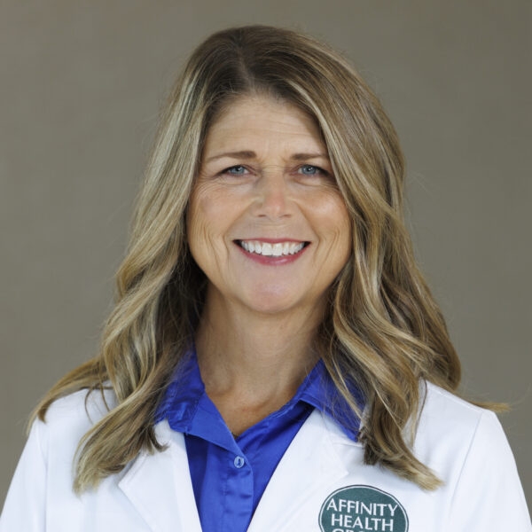 A woman with long, wavy blonde hair wearing a white lab coat and blue shirt, smiling, with an "Affinity Health Group" badge.