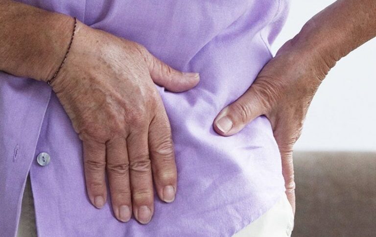 A person in a purple shirt holds their right hip, pressing with both hands, suggesting discomfort or pain in the hip area.