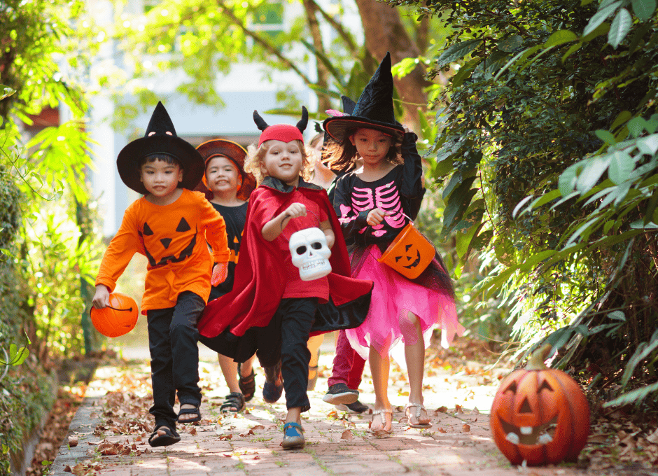 Four children in Halloween costumes, including witches and a devil, run outside on a path with pumpkins and trees around them.