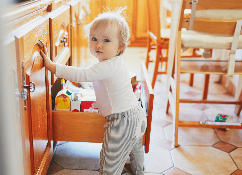 A toddler stands by an open kitchen drawer, holding the handle, with food items visible inside the drawer.