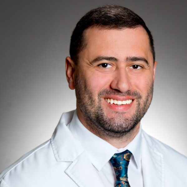 A man in a white medical coat and patterned tie smiles at the camera against a plain gray background.
