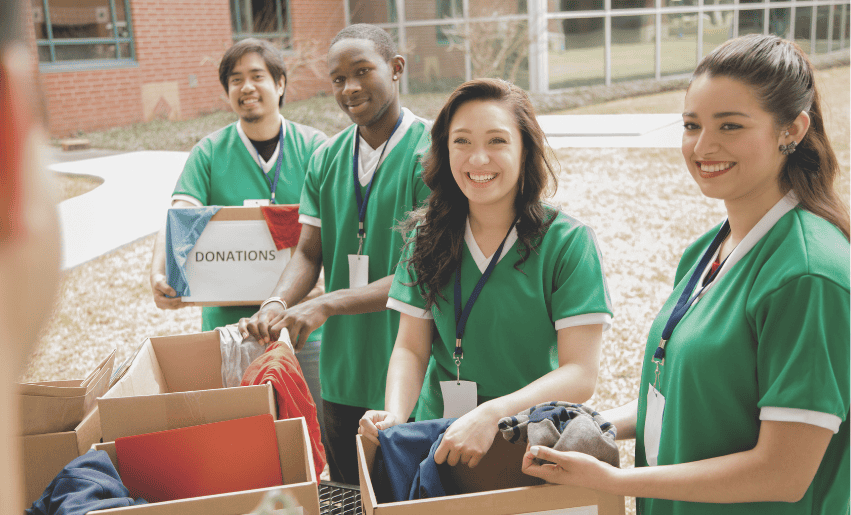 Four people in green shirts smiling and sorting clothes into donation boxes outdoors near a building.