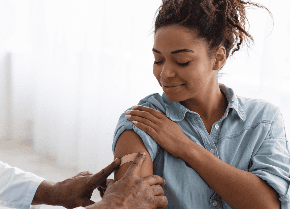 A woman in a denim shirt smiles and looks away as a healthcare professional prepares to give her an injection in her upper arm.