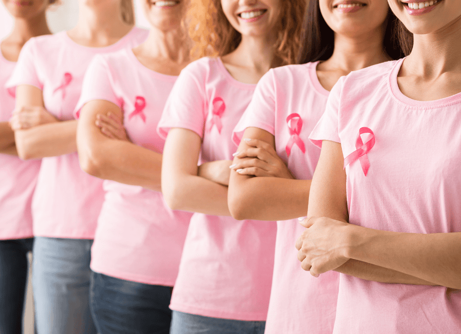 A group of women in pink shirts with pink breast cancer awareness ribbons stand side by side with their arms crossed.