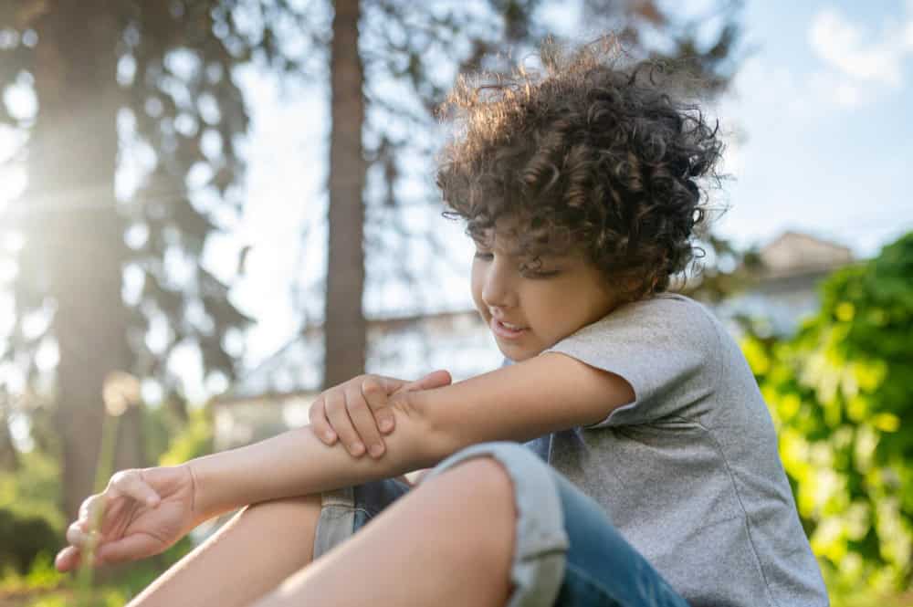 A child with curly hair sits outside, looking at and touching their arm, with greenery and trees in the background.