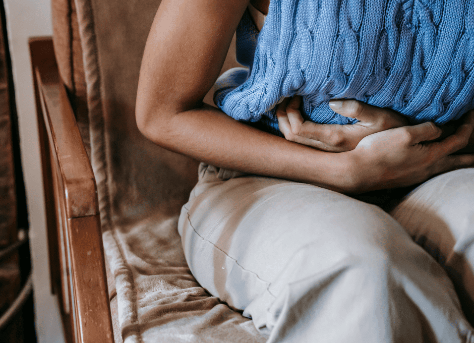 A person sitting on a chair holds their stomach with both hands, wearing a blue sweater and light-colored pants.