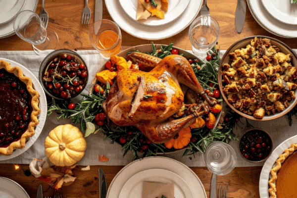 A roasted turkey is surrounded by stuffing, pie, cranberries, and drinks on a decorated wooden table set for a meal.