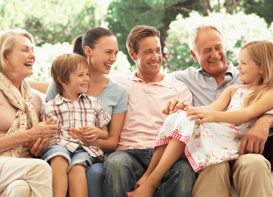 Three adults, two children, and an older woman and man sit together on an outdoor bench, smiling and talking, with greenery in the background.