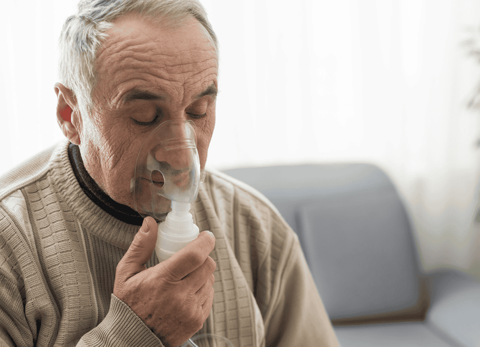 An older man wearing a beige sweater uses a nebulizer mask while sitting indoors, eyes closed.