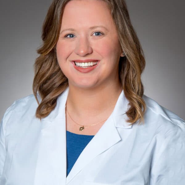 A woman with shoulder-length light brown hair wearing a white lab coat smiles at the camera against a plain gray background.