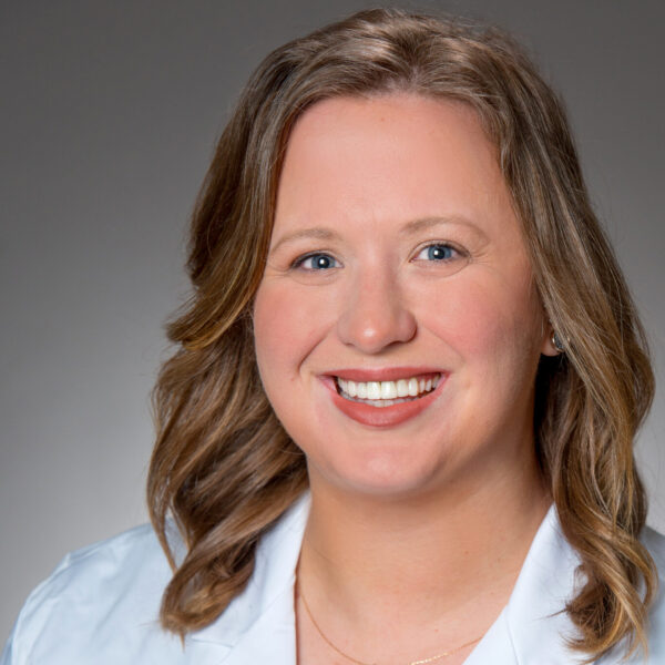 A woman with shoulder-length light brown hair wearing a white lab coat smiles at the camera against a plain gray background.