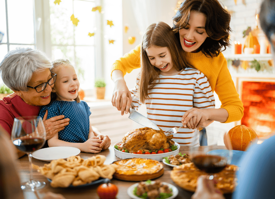 Four people, including two children, gather around a table while a woman carves a roast turkey; various dishes and drinks are on the table.