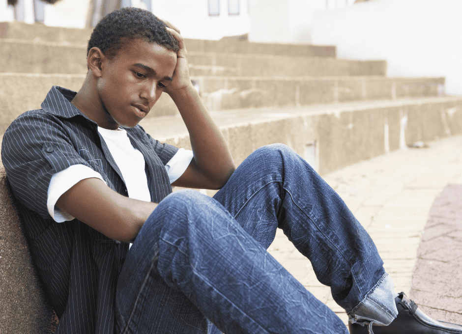 A teenage boy sits on outdoor steps, resting his head on his hand and looking down with a thoughtful expression.