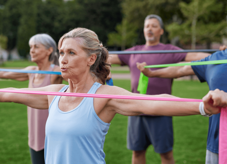 A group of older adults exercise outdoors, stretching resistance bands with arms extended to the sides.