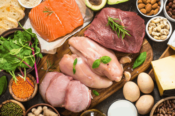 A variety of protein-rich foods, including raw salmon, beef, chicken, eggs, cheese, nuts, seeds, avocado, bread, and leafy greens.