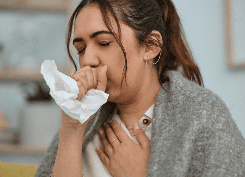A woman wrapped in a blanket holds a tissue and coughs, with one hand on her chest, appearing to feel unwell.