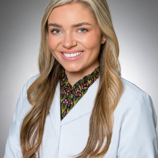 A woman with long blonde hair wears a white lab coat and patterned shirt, smiling at the camera against a neutral background.