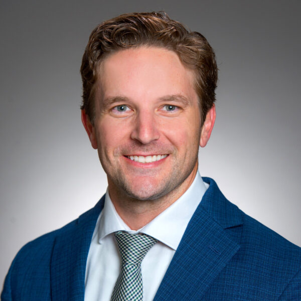 A man in a blue suit, white shirt, and patterned tie smiles in a professional headshot against a plain gray background.