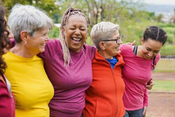 Four women standing outdoors with arms around each other, smiling and laughing together in casual clothing.