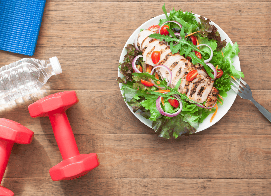A plate of grilled chicken salad with tomatoes, onions, and greens sits beside red dumbbells, a water bottle, and a blue exercise mat.