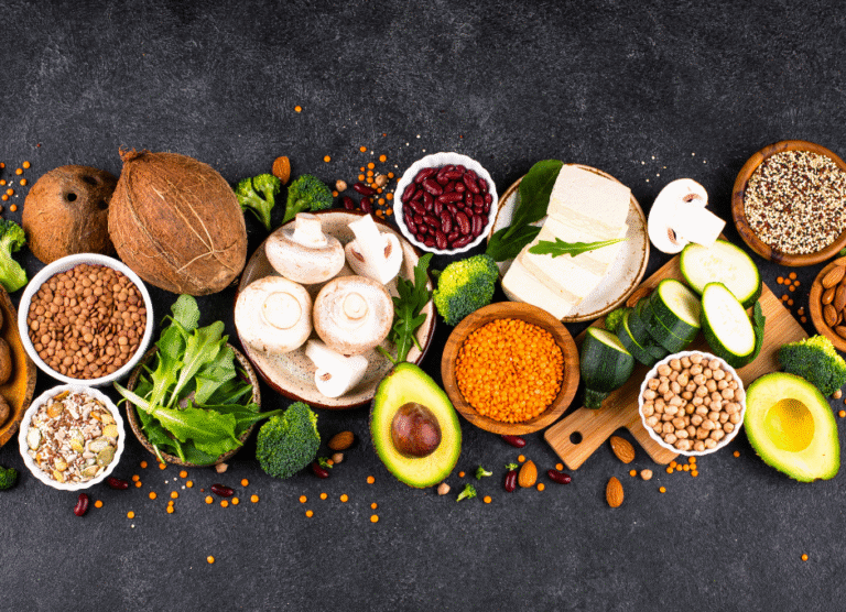 Assorted fresh vegetables, legumes, seeds, tofu, and avocado arranged on a dark surface, viewed from above.